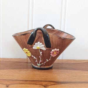 Peut inclure: Un panier en céramique marron avec une poignée noire et des ornements floraux. Le panier présente un motif texturé imitation bois et est orné de fleurs blanches, jaunes et roses. La base est noire.
