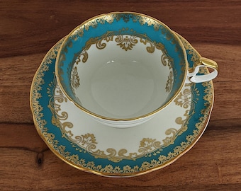 May include: A white teacup and saucer with a teal blue and gold floral design. The cup and saucer have a gold rim.