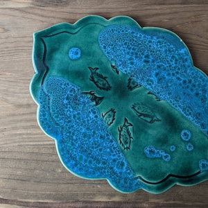 May include: A teal blue ceramic serving dish with a scalloped edge and a design of stylised fish in a bubble pattern.