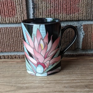 Puede incluir: Taza de cerámica negra con un diseño floral rosa y verde. La taza tiene una asa grande.