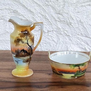 Könnte beinhalten: Ein Vintage-Set aus Porzellan-Kanne und Schüssel mit handbemaltem Landschaftsmotiv. Die Kanne hat eine breite Öffnung und einen geschwungenen Henkel. Die Schüssel ist rund und hat einen goldenen Rand.