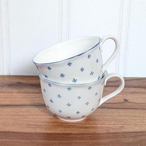 Puede incluir: Dos tazas de cerámica blanca con motivos florales azules apiladas una encima de la otra. Las tazas tienen un diseño simple y clásico.