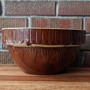 May include: A large brown ceramic mixing bowl with a white rim and a decorative pattern. The bowl has a wide, rounded base and a slightly flared lip.