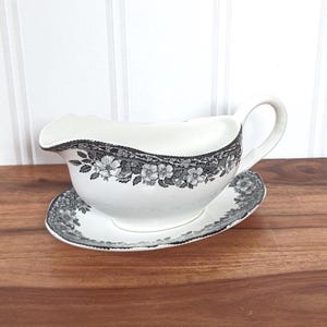 Puede incluir: Salsera blanca con platillo a juego. La salsera tiene un asa y un pico. Ambas piezas están decoradas con un estampado floral en blanco y negro alrededor del borde. El conjunto está colocado sobre una superficie de madera.