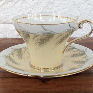 Puede incluir: Juego de taza y platillo de té vintage. La taza y el platillo son de color crema con ribete dorado y un diseño similar al trigo en gris. La taza tiene un asa curvada y un borde festoneado. El platillo tiene un diseño a juego.