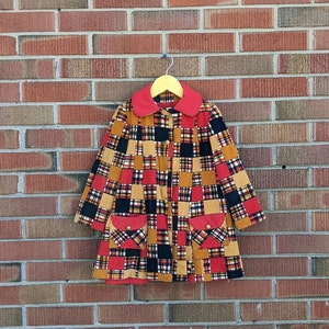 May include: A red, black and brown plaid coat with a red collar and two patch pockets. The coat is hanging on a wooden hanger against a brick wall.