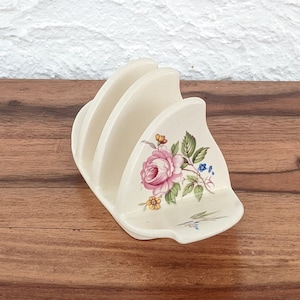 Pode incluir: Um suporte para torradas de cerâmica branca com um design floral que apresenta uma rosa rosa, folhas verdes e flores amarelas e azuis.