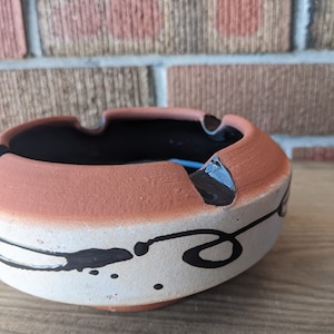 Peut inclure: Un cendrier en terre cuite avec un glaçage blanc et un motif abstrait noir. Le cendrier a un bol large et peu profond et une base arrondie.