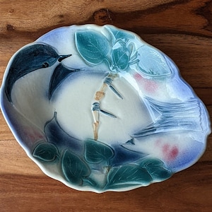 Puede incluir: Un plato de cerámica blanca con un diseño floral azul y verde. El plato presenta un pájaro estilizado con alas azules y blancas y un tallo verde con hojas.