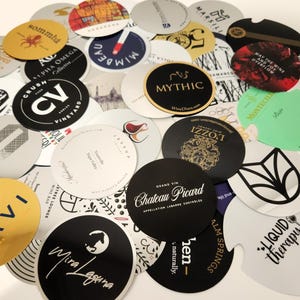 Könnte beinhalten: Eine Sammlung verschiedener Weinkorkuntersetzer mit unterschiedlichen Designs und Logos. Die Untersetzer sind rund und aus Pappe oder einem ähnlichen Material. Einige Untersetzer tragen Texte wie "Chateau Picard" und "Liquid Therapy".