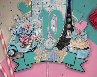 May include: A decorative cake topper featuring the number 10 in teal, a black glitter Eiffel Tower, a blue scooter with floral accents, a cupcake with a patterned bow, and a teal banner with the name Finley in glittery script lettering.