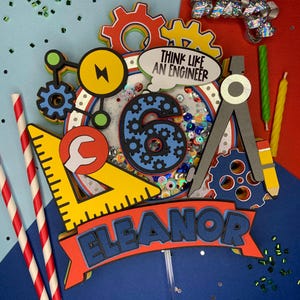 Może przedstawiać: Kolorowy topper na tort z niebieską cyfrą sześć, kołami zębatymi, kątomierzem, kluczem, błyskawicą i napisami "Think Like An Engineer" i "Eleanor".