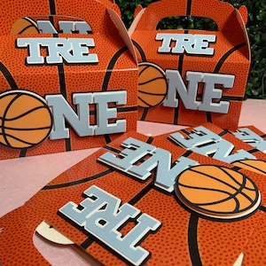 Op de afbeelding: Oranje en zwarte basketbal-thema favoriete dozen met een blauwe "ONE" en "TRE" op elke doos. De dozen hebben een handvat en zijn perfect voor een basketbal-thema feest.