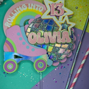 May include: A pink and blue roller skate cake topper with a disco ball, rainbow, and the words "Rolling into 13" and "Olivia".