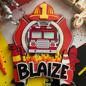 May include: A layered paper cake topper featuring a red fire truck, a firefighter's helmet with the number 1, and the name "BLAIZE" in black and white. Includes a fire extinguisher, fire hydrant, and flames.
