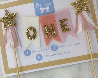 Puede incluir: Un adorno para pastel con una estrella dorada con purpurina y una pancarta que dice "ONE" en letras doradas con purpurina. La pancarta está hecha de fieltro rosa, blanco y melocotón. La estrella está en un palo de madera.
