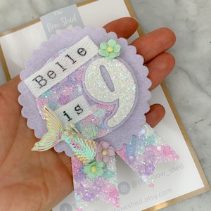 May include: Purple felt birthday badge with a white banner that says "Belle is 9". The number 9 is covered in iridescent glitter. A mermaid tail with iridescent scales is attached to the badge. The badge is decorated with pastel flowers.