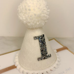 May include: White felt party hat with a silver glitter number one and a white pom pom on top. The hat is attached to a headband.