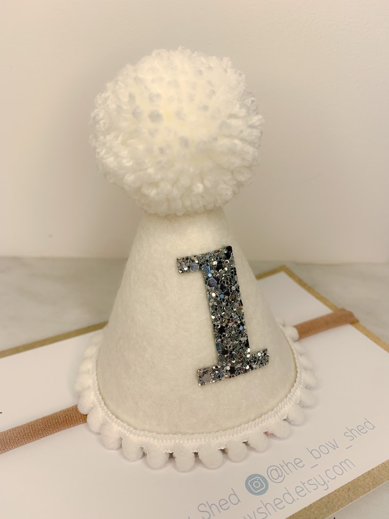 May include: White felt party hat with a silver glitter number one and a white pom pom on top. The hat is attached to a headband.