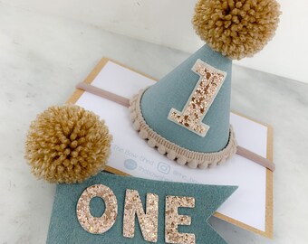 May include: A dusty blue party hat with a beige pom-pom, a glittery number one, and a beige pom-pom trim, paired with a matching dusty blue felt flag on a wooden stick featuring the glittery text ONE, displayed on a branded card.