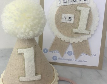 May include: A beige felt party hat with a white felt number one and a white pom-pom on top. A matching beige felt badge with a white felt number one and the text "Indie is 1" in black felt.