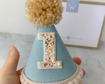 May include: A light blue felt party hat with a gold glitter number one on the front. The hat has a white pom-pom on top and a white pom-pom trim around the bottom.