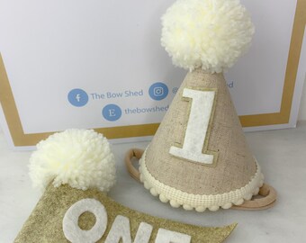May include: A beige felt banner with the word "ONE" in white felt letters. A beige felt party hat with a white pom-pom on top and a white felt number "1" on the front. The hat has a white elastic band.