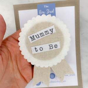 Peut inclure: Un badge en feutre beige avec des bords festonnés blancs et un ruban blanc. Le badge dit "Mummy to Be" en broderie noire.