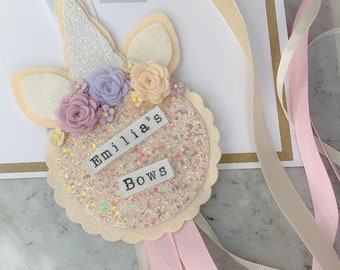 May include: A white felt unicorn head with pink, purple and yellow felt flowers. The unicorn head has a sparkly pink and white circle with the text "Emilia's Bows" on it. The unicorn head is attached to a pink and white ribbon.
