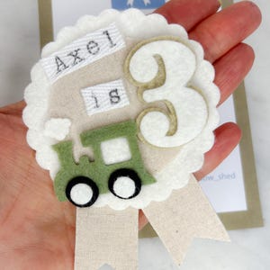 May include: A handmade birthday badge with a white scalloped edge. The badge features the name "Axel", the number "3", and a green felt train. A beige ribbon extends from the bottom.