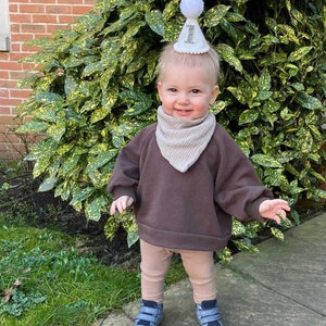 May include: A toddler wearing a brown sweatshirt, tan leggings, and blue shoes. The toddler is wearing a white party hat with the number "1" on it. The toddler is standing in front of a green bush.