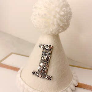 May include: White felt party hat with a silver glitter number one and a white pom pom on top. The hat has a white fuzzy trim around the bottom.