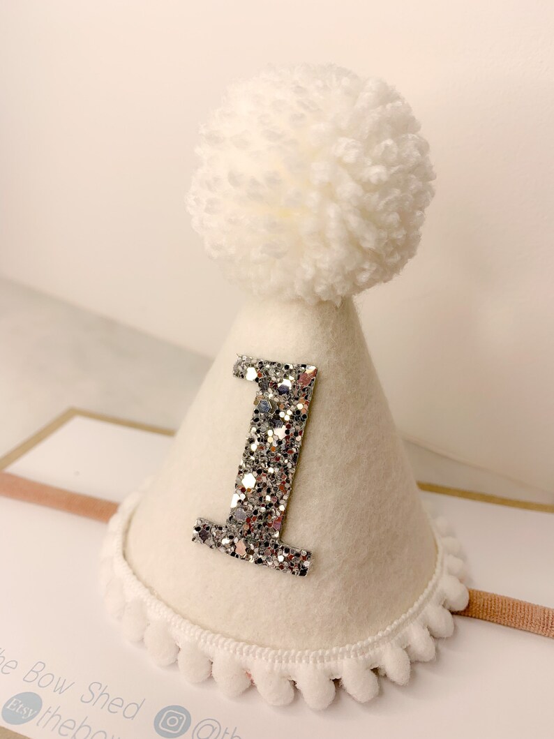 May include: White felt party hat with a silver glitter number one and a white pom pom on top. The hat has a white fuzzy trim around the bottom.