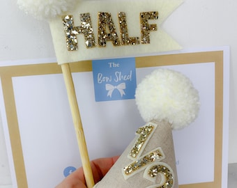 May include: A beige felt party hat with a white pom-pom and a gold glitter 1/2 appliqué, paired with a matching beige felt flag on a wooden stick featuring the word HALF in gold glitter letters and a white pom-pom on the corner.
