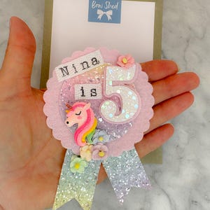 May include: A circular felt badge featuring a glittery rainbow background, a pink unicorn figure, and a large white glittery number 5. The text Nina is is printed on a white fabric strip. Two glittery ribbon tails hang below with small floral accents.