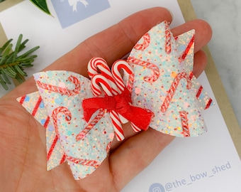 May include: A white and red glitter bow with a candy cane print and three red and white candy canes in the center. The bow is on a hand holding a card with the text "The Bow Shed" and social media information.