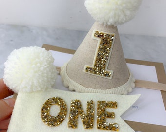 May include: A white felt banner with gold glitter letters spelling "ONE" on a wooden stick. A white felt party hat with a gold glitter "1" and a white pom-pom on top.