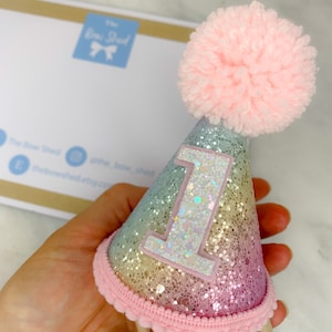 May include: A rainbow glitter party hat with a pink pom pom on top and a white number one on the front. The hat is attached to a headband.