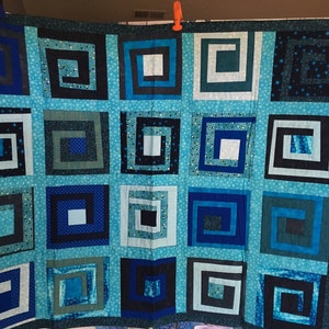 Puede incluir: Una colcha con un patrón geométrico de cuadrados dentro de cuadrados. La colcha está hecha de telas azules, verde azulado y negras.