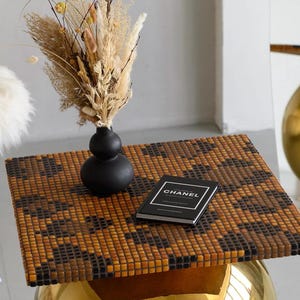 Könnte beinhalten: Ein dekorativer Tisch mit einer Mosaikfliesenplatte in Braun- und Schwarztönen. Eine kleine schwarze Vase enthält getrocknete Blumen. Ein Buch mit dem Wort "CHANEL" liegt auf dem Tisch. Der Tisch hat ein goldfarbenes, abgerundetes Gestell.
