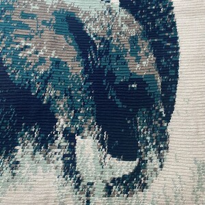 May include: A close-up of a crocheted tapestry featuring a blue and green abstract design. The design resembles a stylized animal or creature.