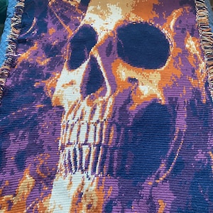 May include: A crocheted throw blanket with a skull design. The skull is orange and white, and the background is purple and blue.