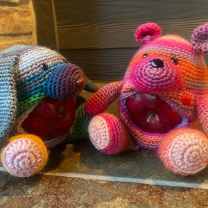 Op de afbeelding: Twee gehaakte knuffels, een beer en een hond, beide met appels in hun lichaam. De beer is roze, oranje en rood, terwijl de hond blauw, groen en paars is.