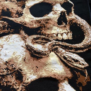 Könnte beinhalten: Schwarzer Stoff mit einem detaillierten Design aus zwei Totenköpfen und einer Schlange. Die Totenköpfe sind in Braun- und Beigetönen gehalten, wobei sich die Schlange um sie windet. Der Hintergrund ist einfarbig schwarz und erzeugt einen auffälligen Kontrast.