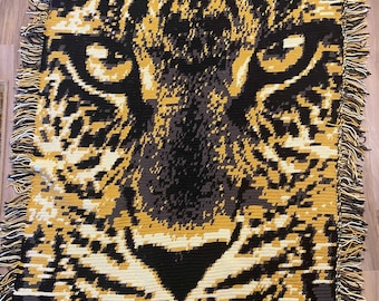 May include: A woven tapestry featuring a close-up of a leopard's face. The tapestry is made of yarn in shades of black, brown, and gold. The leopard's eyes are a bright green.