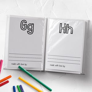 May include: An open plastic-bound activity book featuring the letters Gg and Hh on separate pages, each with three blank writing lines and the printed text Made with love by. Several colorful felt-tip markers are scattered around the book.