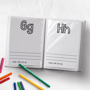May include: A white book with black lines and the letters "Gg" and "Hh" printed on the pages. The book is open and there are colorful markers on a white surface next to it.