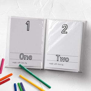 May include: A white book with two pages, each with a black outline of a number and the word for the number. The first page has the number "1" and the word "One". The second page has the number "2" and the word "Two". The book has the text "Made with love by" on each page. There are colorful markers on a white surface.