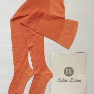 May include: A pair of orange tights with a textured pattern, laid out on a white surface. The tights are accompanied by a white paper package with the text "Bi Bi Calze Luana" in gray.