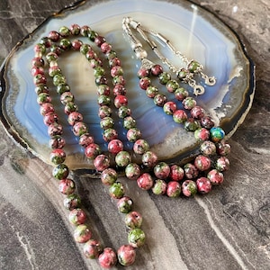 May include: A prayer bead mala made with green and pink speckled beads. The mala is laid out on a gray and white agate slab.